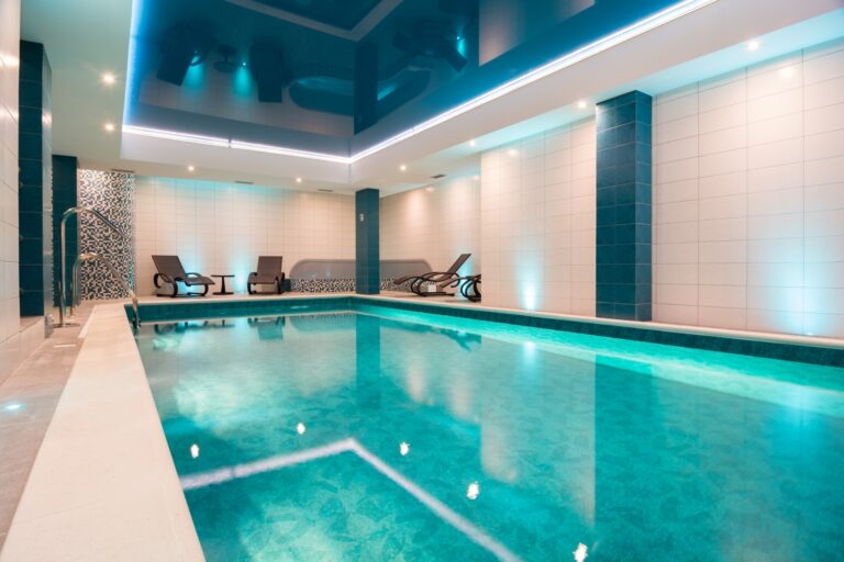 Kryty basen hotelowy z rzędem leżanek pod ścianą, podświetlany na niebiesko i turkusowo