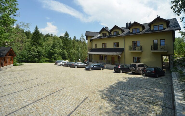 Dom pod Koziołkami – widok od strony brukowanego parkingu na żółty budynek pensjonatu wśród lasu