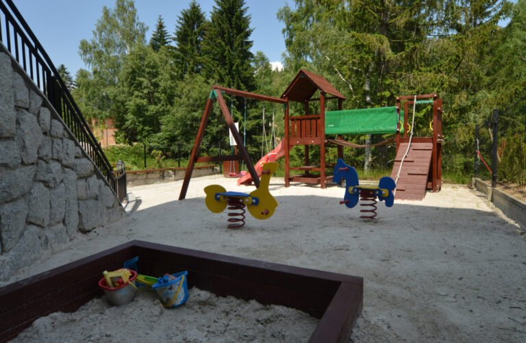 Plac zabaw z piaskownicą, bujakami na sprężynach i drewnianym zestawem wspinaczkowym wśród świerków